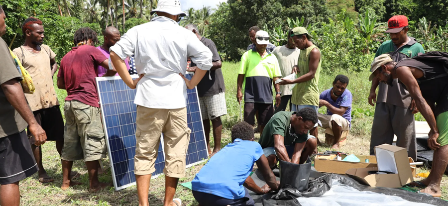 MakiraGold - CERES Fair Food A diverse group of men gathers outdoors around a large solar panel, assembling a solar power kit on the ground.