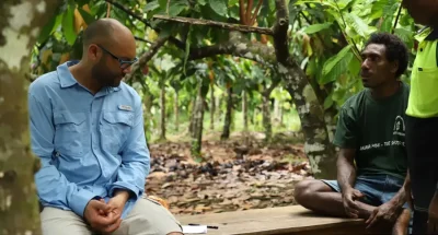 MakiraGold2 - CERES Fair Food - CERES Fair Food Two men sit and chat on a wooden bench in a Solomon Islands cacao orchard, one in a light blue shirt and glasses, another in a dark green T-shirt; a third person stands nearby in a high-visibility shirt.
